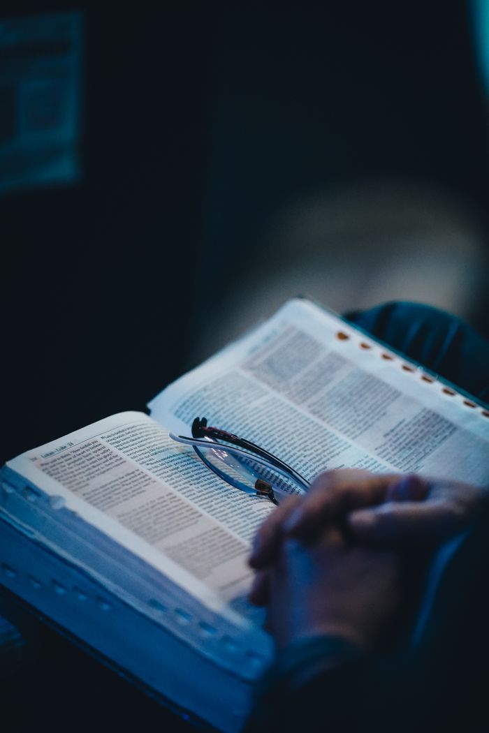 A person holding an open book with glasses resting on the pages, depicting quiet reading time indoors.