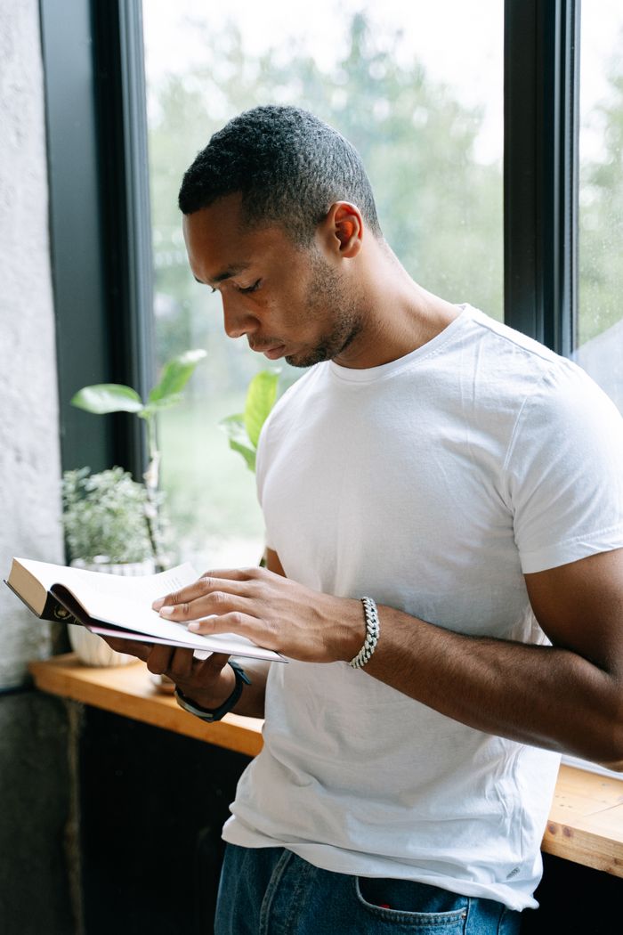 A focused man reads a book by the window with indoor plants in the background.